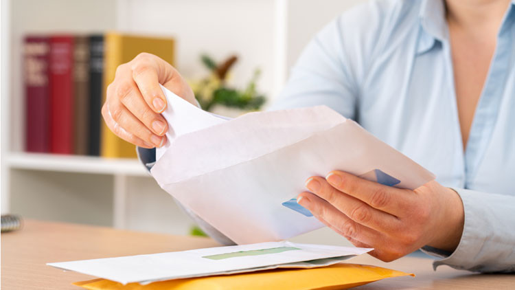 A woman sits at a table putting a letter into an envelope. There are two other envelopes on the table.