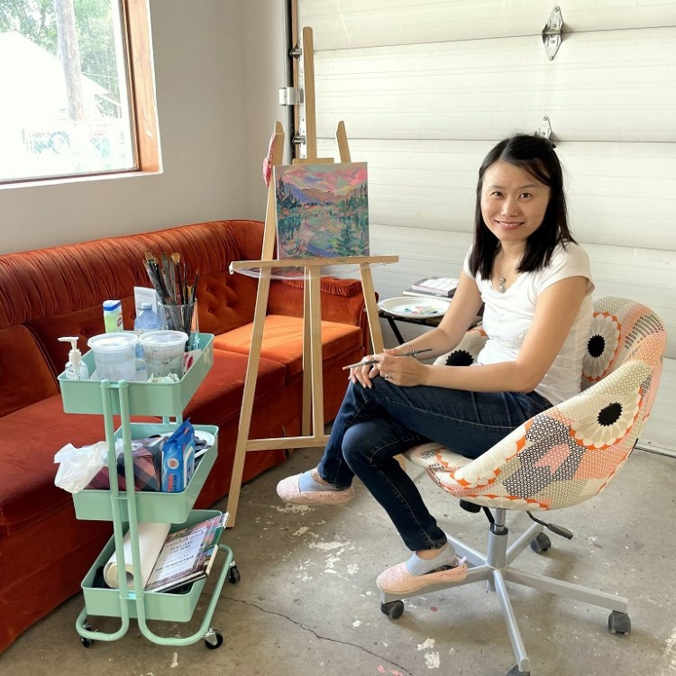 Aeris sits in a chair next to her artwork and smiles.