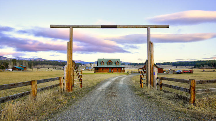 Rural driveway with a wooden entrance gate and a red barn house in the distance