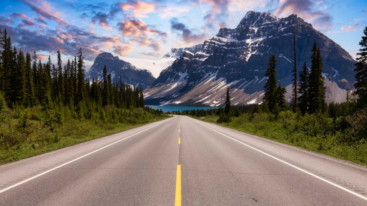 A single lane road heads towards the mountains. The sun is setting behind the mountains and sky is scattered with pink and purple clouds.