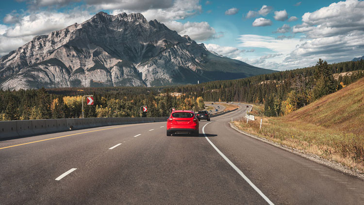 Cars driving along a winding highway in the mountains under a partially-cloudy sky.